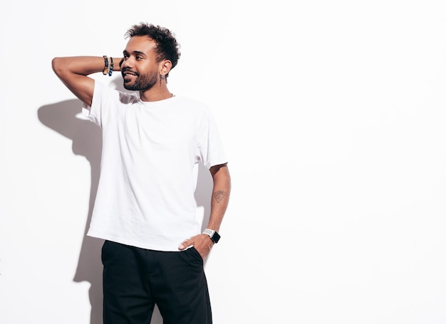 Beau modèle hipster souriant Sexy homme mal rasé vêtu de vêtements d'été t-shirt et jeans Mode homme posant près d'un mur blanc en studio à la journée ensoleillée Isolé