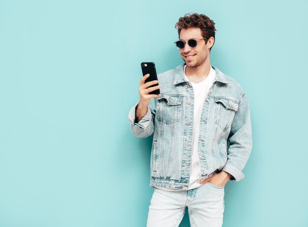 Beau modèle lambersexuel hipster élégant et confiant avec une coiffure frisée Homme sexy vêtu d'une veste en jean Homme de mode tenant un smartphone Regardant l'écran du téléphone portable