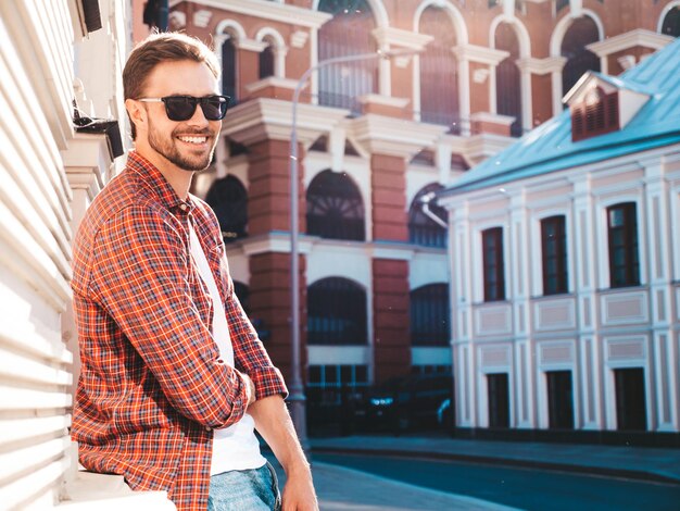 Beau modèle lambersexuel hipster élégant et souriant. Homme moderne vêtu d'une chemise à carreaux rouge. Homme de mode posant près de gratte-ciel sur le fond de la rue en lunettes de soleil. À l'extérieur au coucher du soleil