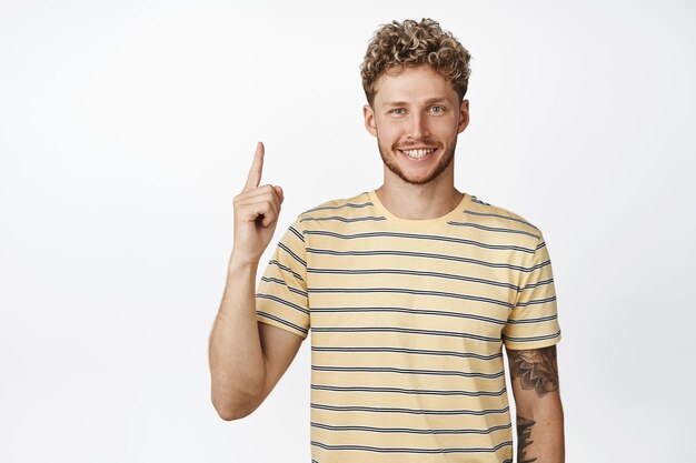 Beau modèle masculin caucasien aux cheveux blonds et à la barbe pointant le doigt vers le haut et souriant montrant l'annonce démontrant la bannière de vente vers le haut sur fond blanc