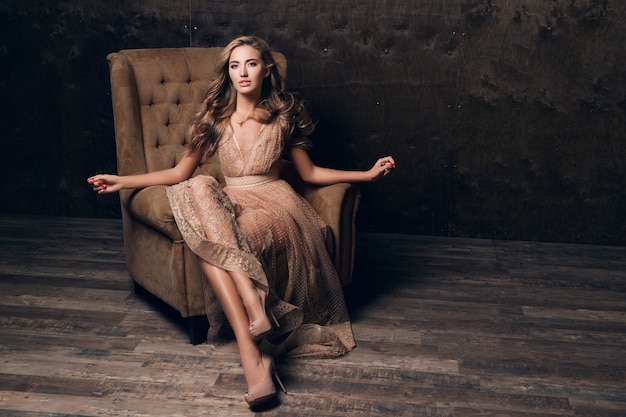 Beau modèle sexy femme en robe de soirée en dentelle brillante posant assis dans une chaise de couleur beige