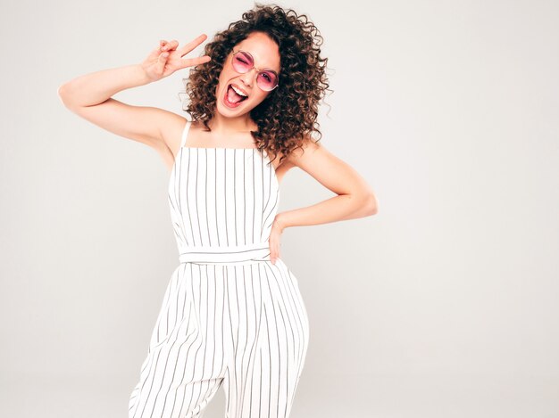 Beau modèle souriant avec coiffure afro curls vêtu de vêtements d'été hipster.Tendance femme drôle à lunettes de soleil.Affiche le signe de la paix