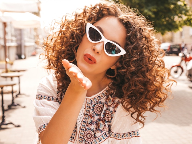 Beau modèle souriant avec une coiffure de boucles afro habillée en robe blanche d'été hipster.