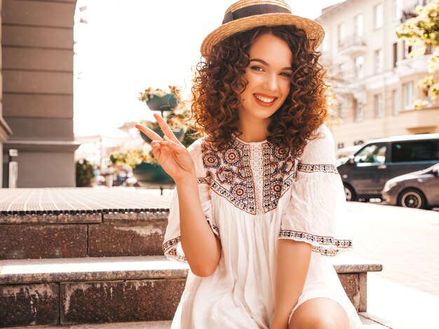 Beau modèle souriant avec une coiffure de boucles afro habillée en robe blanche d'été hipster.