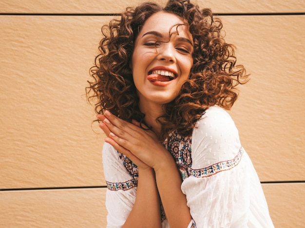 Beau modèle souriant avec une coiffure de boucles afro habillée en robe blanche d'été hipster.