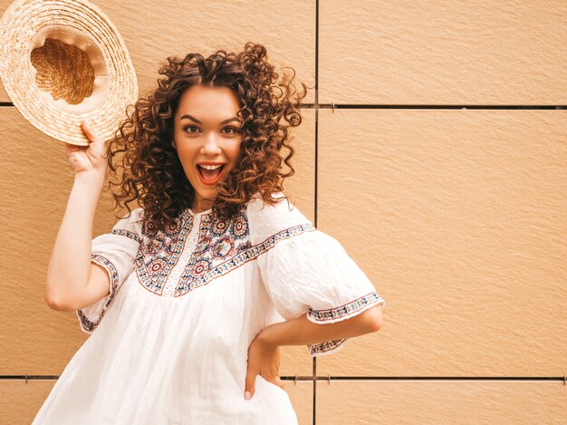 Beau modèle souriant avec une coiffure de boucles afro habillée en robe blanche d'été hipster.