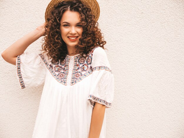 Beau modèle souriant avec une coiffure de boucles afro habillée en robe blanche d'été hipster.