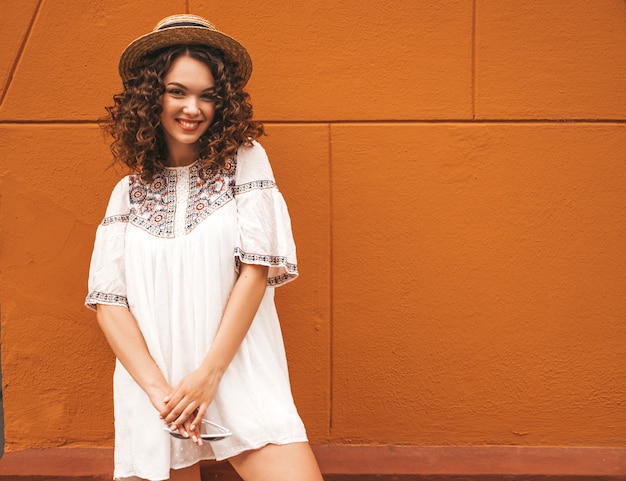 Beau modèle souriant avec une coiffure de boucles afro habillée en robe blanche d'été hipster.