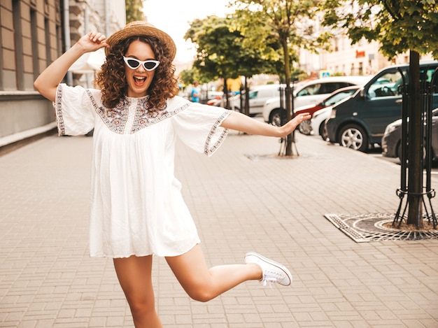 Beau modèle souriant avec une coiffure de boucles afro habillée en robe blanche d'été hipster.