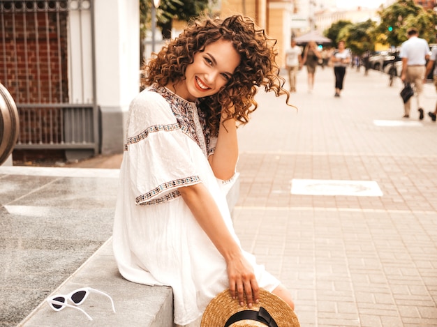 Beau modèle souriant avec une coiffure de boucles afro habillée en robe blanche d'été hipster.