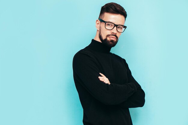 Beau modèle souriant Sexy homme élégant vêtu d'un pull à col roulé noir et d'un jean Mode hipster mâle posant près du mur bleu en studio Isolé dans des lunettes ou des lunettes Bras croisés