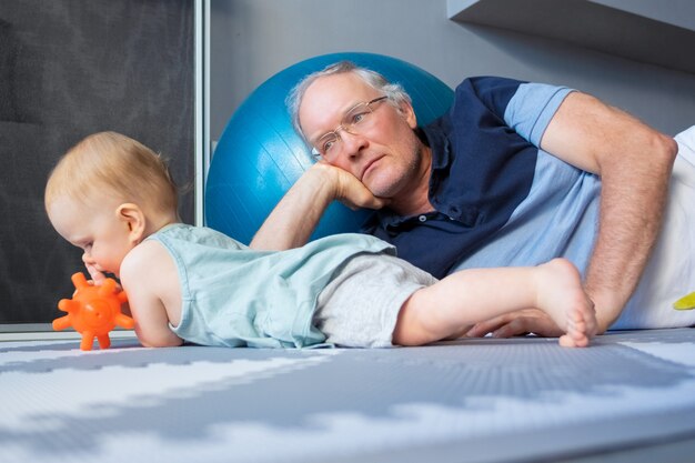 Beau nouveau-né aux cheveux roux allongé sur le ventre et jouant avec un jouet. Bébé sur un sol mou. Grand-père sérieux à lunettes couché près de petit-enfant et regardant bébé. Concept de famille, petite enfance et enfance