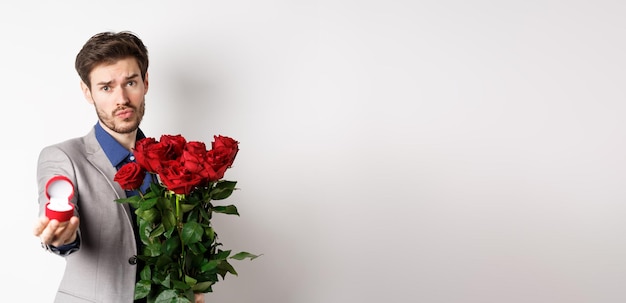 Photo gratuite beau petit ami en costume demandant de l'épouser debout avec bouquet de roses rouges et rin de fiançailles