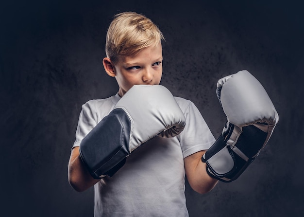 Un beau petit garçon boxeur aux cheveux blonds vêtu d'un t-shirt blanc avec des gants prêts à se battre. Isolé sur le fond texturé sombre.