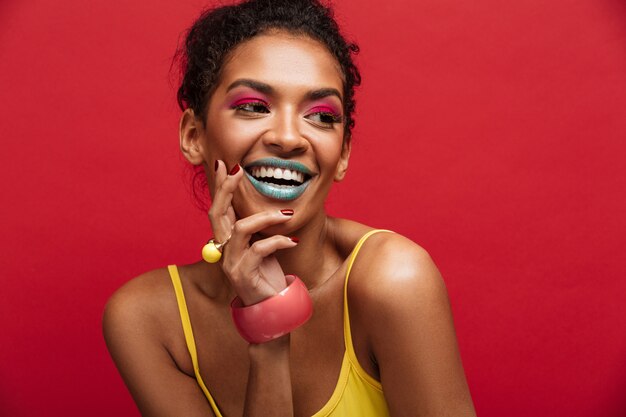 Beau portrait de modèle féminin afro-américain heureux en chemise jaune souriant et posant sur l'appareil photo, isolé sur mur rouge