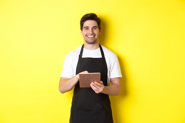 Photo gratuite beau serveur prenant des commandes, tenant une tablette numérique et souriant, portant un uniforme de tablier noir, debout sur fond jaune.