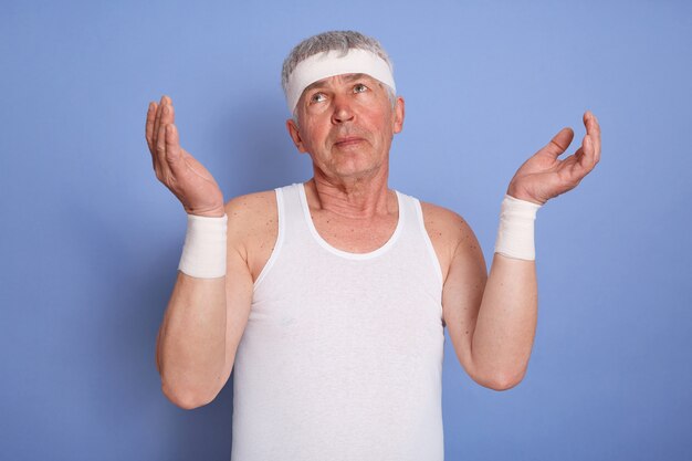 Beau sportif d'âge moyen, a l'air désemparé et confus, garde les bras levés, n'a aucune idée, porte un t-shirt blanc sans manches et entend un groupe, debout isolé.