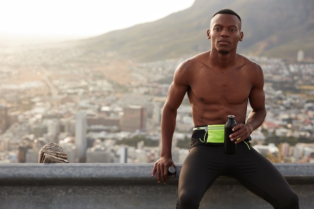 Beau sportif au hiar foncé, prend une pause après un entraînement intensif en montagne, tient une bouteille d'eau fraîche, a soif, respire l'air frais plein d'énergie. Concept de sport et de motivation