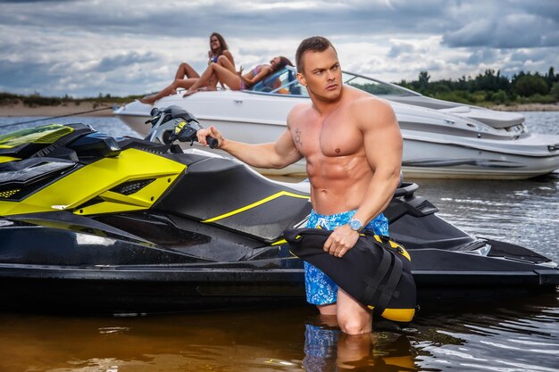 Un beau sportif torse nu avec un corps musclé debout près de jet ski sur un littoral.