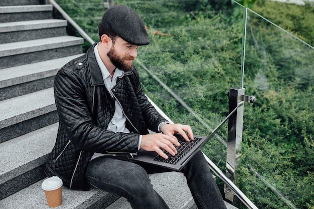 Beau Travailleur Heureux Buvant Du Café Et Travaillant Sur Un Ordinateur Portable Assis à L'extérieur Dans Les Escaliers