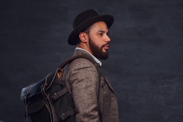 Photo gratuite beau voyageur afro-américain dans une veste marron et un chapeau avec le sac à dos, se tient dans un studio. isolé sur un fond sombre.