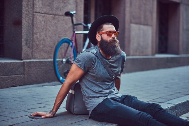 Beau voyageur hipster avec une barbe élégante et un tatouage sur les bras vêtu de vêtements décontractés avec un sac, assis sur le trottoir, se reposant après une balade à vélo.