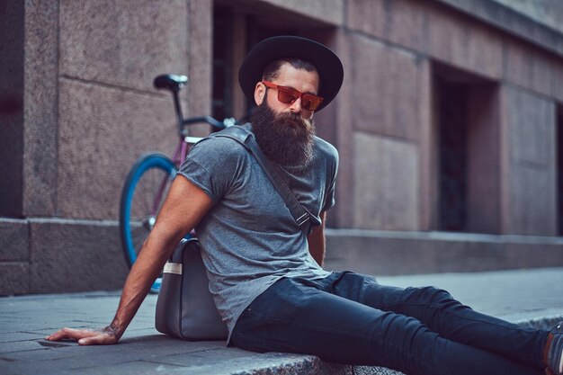 Beau voyageur hipster avec une barbe élégante et un tatouage sur les bras vêtu de vêtements décontractés avec un sac, assis sur le trottoir, se reposant après une balade à vélo.