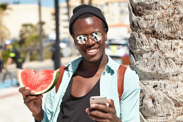 Beau voyageur noir insouciant dans des vêtements urbains élégants posant pour selfie, debout à l'extérieur avec une tranche de pastèque, appuyé en arrière sur un palmier, l'écran du téléphone se reflète dans ses abat-jour en miroir