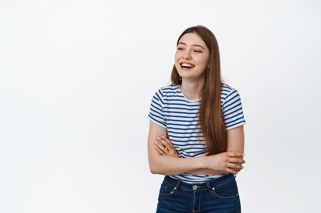 Beauté et art de vivre. Heureuse jeune femme riant, souriante et regardant de côté quelqu'un, debout les bras croisés en tenue décontractée sur blanc.