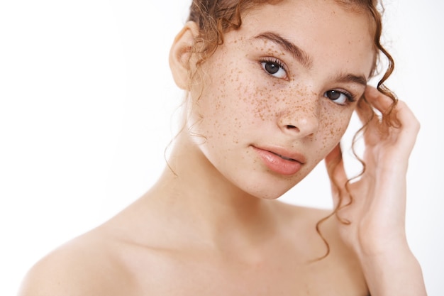 Photo gratuite beauté, concept de soins de la peau. tendres taches de rousseur nues de jeune femme rousse regardent la caméra sensuellement toucher doucement la peau du visage pure, propre et non altérée en appliquant des produits de cosmétologie de traitement quotidien après la douche