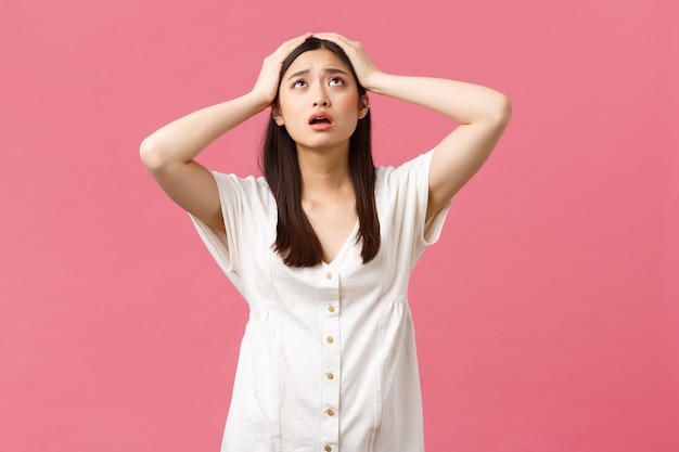 Beauté, émotions des gens et concept de loisirs d'été. Jeune femme asiatique désespérée et frustrée en panique, regarde le ciel, aide de dieu, saisis la tête alarmée, debout sur fond rose anxieux.