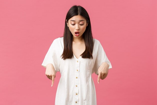 Beauté, émotions des gens et concept de loisirs et de vacances d'été. Fille asiatique kawaii surprise et excitée en robe blanche, pointant et regardant vers le bas avec un visage heureux amusé, fond rose.