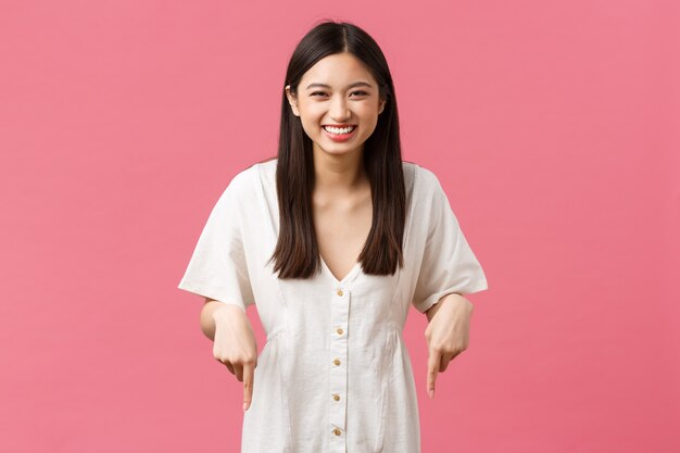 Beauté, émotions des gens et concept de loisirs et de vacances d'été. Fille asiatique mignonne sortante insouciante en robe blanche riant de joie, pointant les doigts vers le bas, invitant à faire la fête ou un événement.