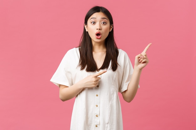 Beauté, émotions des gens et concept de loisirs et de vacances d'été. Jolie fille asiatique impressionnée pointant les doigts dans le coin supérieur droit avec la mâchoire tombante et le visage surpris, debout sur fond rose