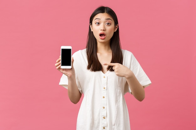 Beauté, émotions humaines et concept technologique. Fille coréenne assez élégante montrant l'application sur l'écran du smartphone. Une femme a l'air choquée en racontant de grandes nouvelles et en pointant du doigt l'application pour téléphone portable.