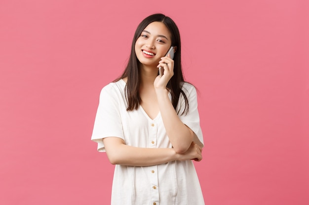 Beauté, émotions humaines et concept technologique. Heureuse fille asiatique en robe blanche parlant au téléphone, tenant un appareil photo mobile et regardant, appelant un ami, fond rose.