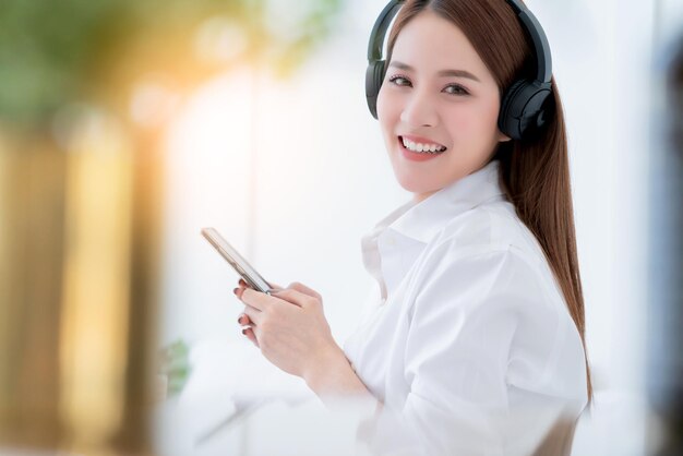 Beauté femme asiatique robe blanche cheveux longs porter un casque et utiliser un smartphone de recherche de type sur un lit blanc avec fond intérieur de chambre blanche palourde et joyeuse