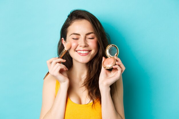 Beauté. Gros plan sur une belle jeune femme ferme les yeux et sourit tout en appliquant des fards à joues sur les joues avec un pinceau de maquillage, debout heureux sur fond bleu