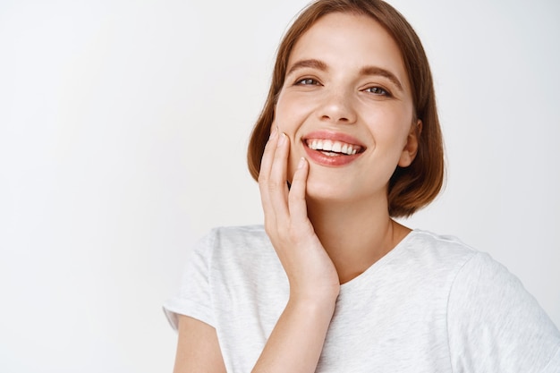 Beauté. Gros plan d'une femme heureuse aux cheveux courts, touchant une peau du visage propre et fraîche et souriante, montrant des dents et un visage parfaits, debout contre un mur blanc