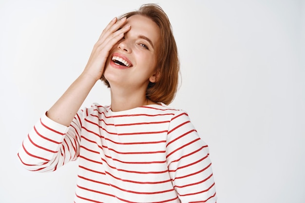 Beauté Heureuse jolie femme aux cheveux courts couvrant la moitié du visage et riant avec des émotions joyeuses candides debout sur fond blanc