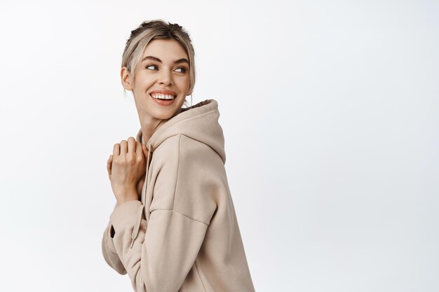 Beauté jeune femme blonde souriante à la recherche de plaisir derrière son épaule lisant l'espace de copie de logo debout dans un sweat à capuche beige sur fond blanc