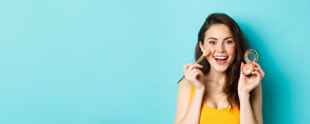 Beauté jeune femme séduisante souriante appliquant du maquillage avec une brosse montrant des rougeurs à la caméra debout