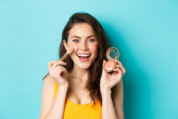 Beauté. Jeune femme séduisante souriante, appliquant le maquillage avec un pinceau, montrant des rougeurs à la caméra, debout sur fond bleu. Espace de copie