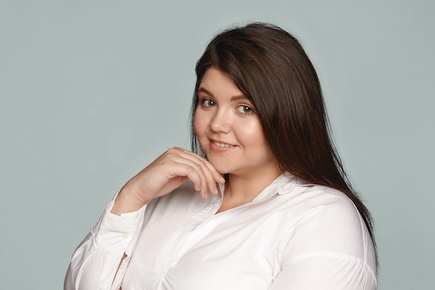 Beauté, positivité corporelle, style, mode et féminité. Plus la taille de la jeune femme en surpoids avec une peau parfaite, des cheveux nets et un sourire joyeux posant, touchant son menton, vêtu d'une chemise blanche