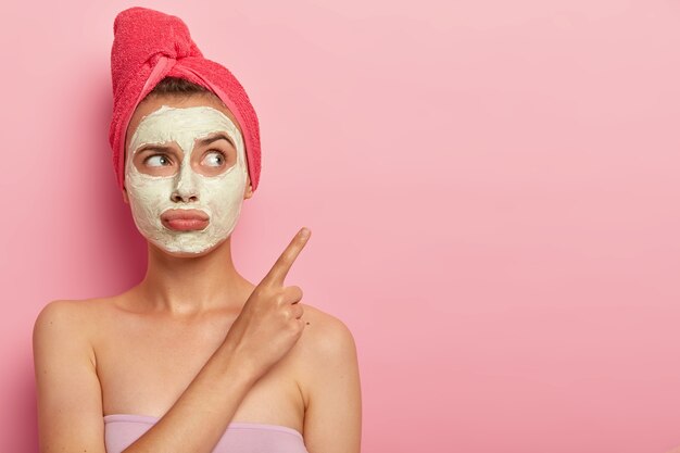 Beauté, spa, concept de traitement. Une femme insatisfaite malheureuse porte un sac à lèvres, applique un masque d'argile pour le rajeunissement, enveloppé dans une serviette de bain, pointe sur l'espace de copie contre le mur rose, veut un résultat rapide