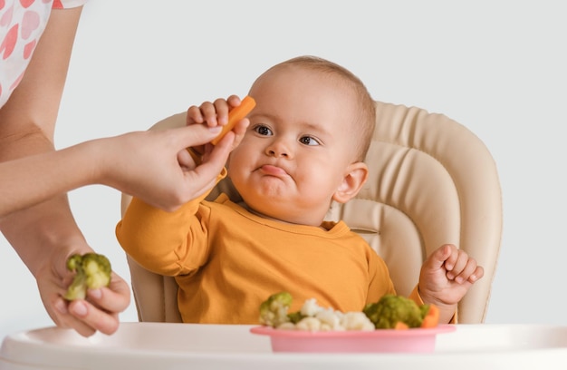 Un Bebe Age De 11 A 15 Mois Prend Des Carottes Bouillies Des Mains De Sa Mere Bebe Sur Une Chaise D Alimentation Manger Des Legumes Bouillis Fond Blanc Photo Premium