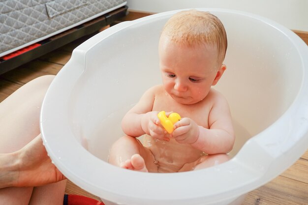 Bébé mignon jouant avec un canard jouet en caoutchouc tout en ayant une baignoire à la maison. Mère baignant le petit enfant dans la baignoire à l'intérieur de la maison. Photo recadrée. Concept de garde d'enfants ou de soins de santé