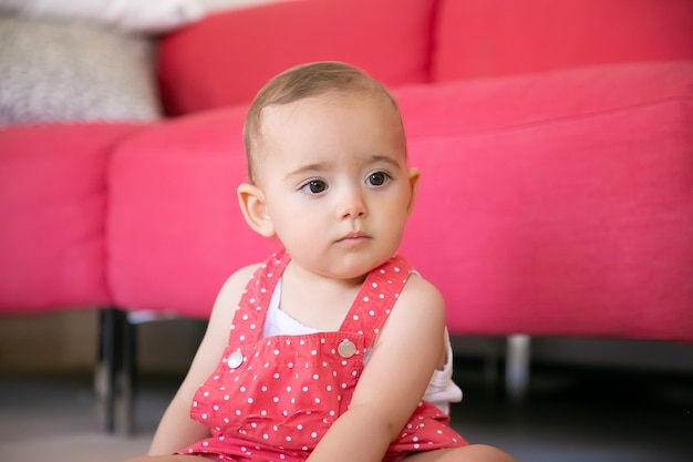 Bébé pensif assis sur le sol dans le salon et penser à quelque chose. Sérieuse petite fille en short salopette rouge à la recherche de suite et assis près du canapé. Concept de week-end, enfance et être à la maison