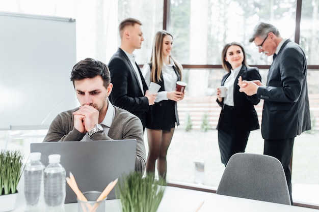 Bel entrepreneur avec ordinateur portable assis dans un bureau moderne et pensant, tandis que ses collègues font une pause avec un café.