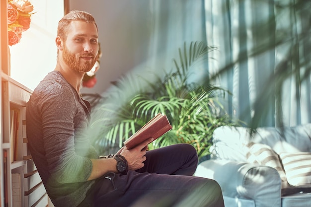 Un bel étudiant rousse élégant en vêtements décontractés, assis sur une chaise dans une maison et tient un livre, regardant une caméra.